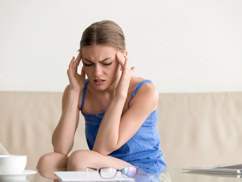 A woman at her desk with both hands pressed to her temples, laptop still open in front of her